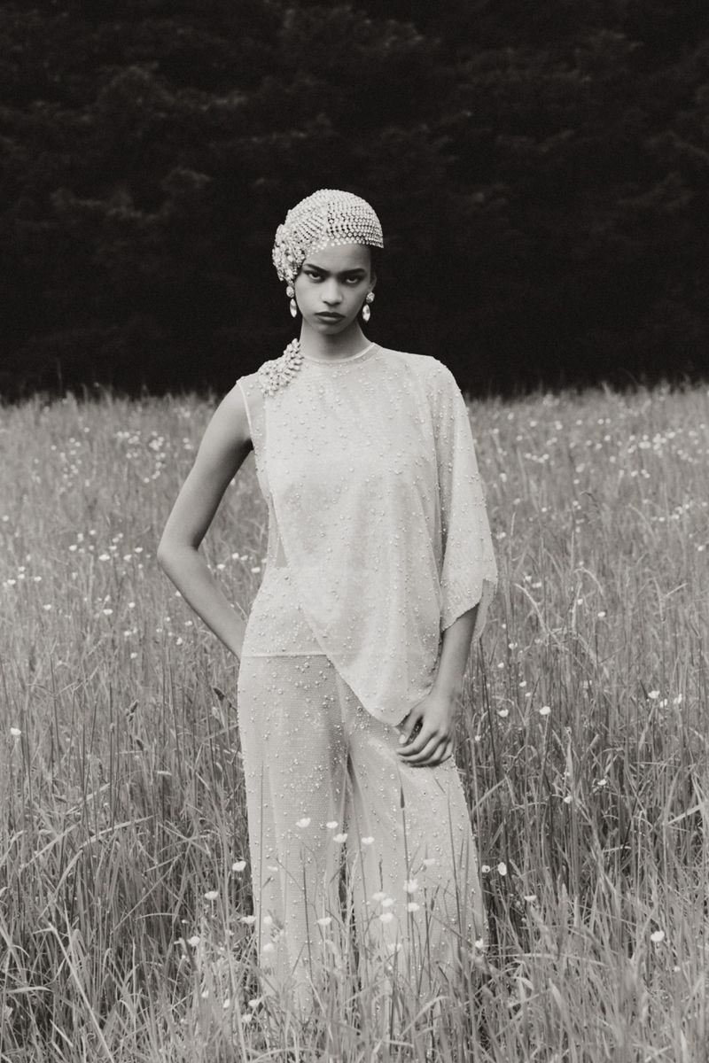 Monochrome portrait of a woman in pearl headpiece and dress standing in a grassy field with flowers, against a dark forest backdrop.