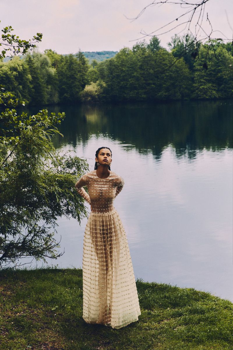 A woman in a long dress stands near a lake surrounded by trees. She is looking up and the scene is captured in soft, natural light.
