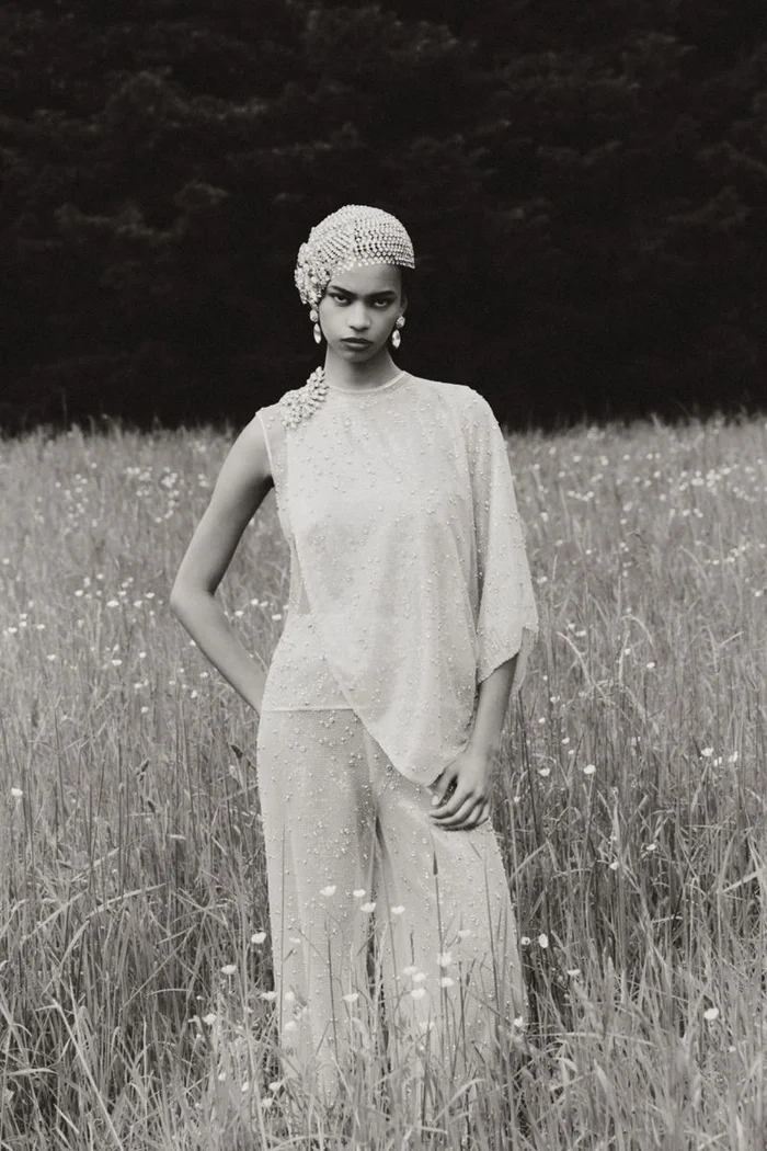 A model wearing beaded haute couture clothing and a matching headpiece standing in a grassy meadow.