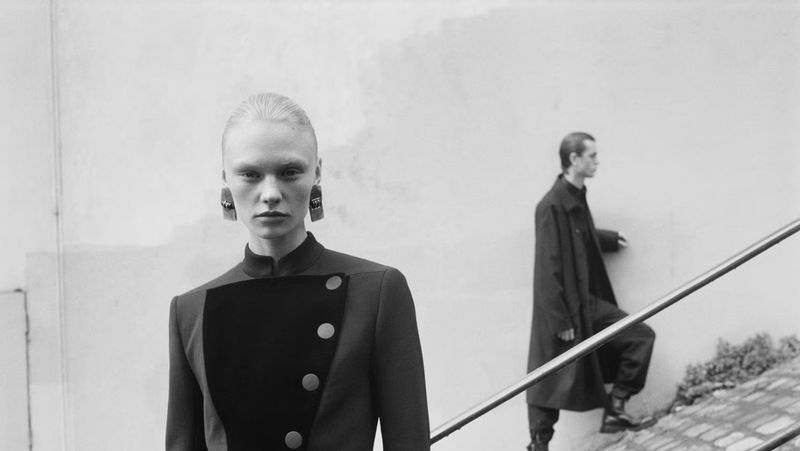 Black-and-white fashion shot featuring a woman in a dark jacket with a man climbing stairs in the background, creating a stylish, moody scene.