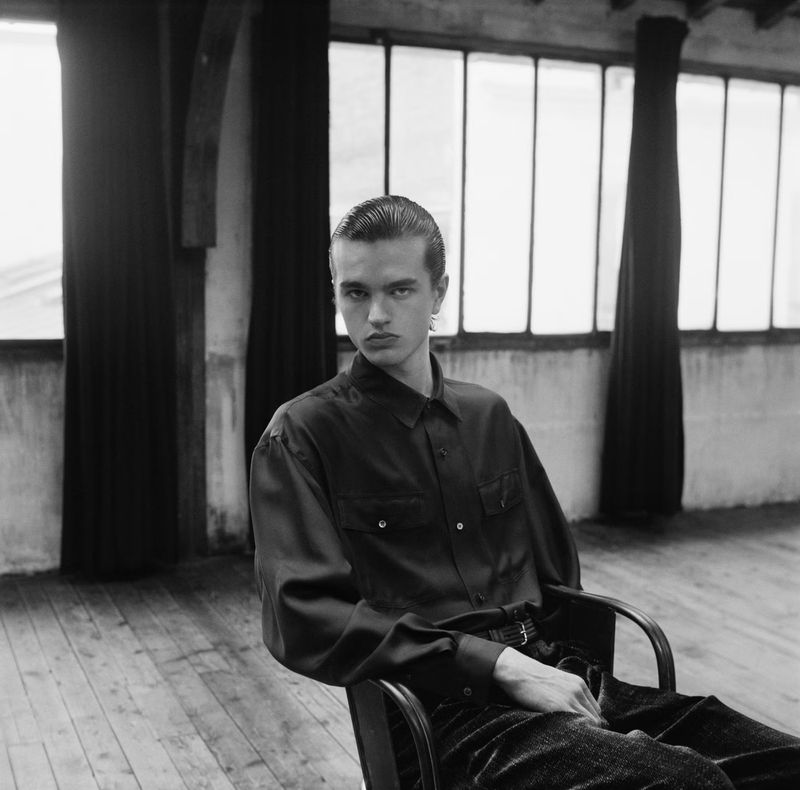 Monochrome portrait of a young adult man with slicked-back hair in a black shirt, sitting in a chair indoors with window light.