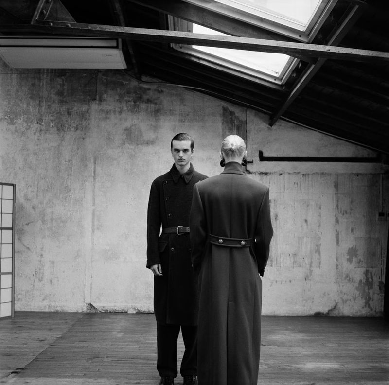 Monochrome image of two men in dark overcoats, one facing forward, one with his back to the viewer, in a spacious room with skylights.