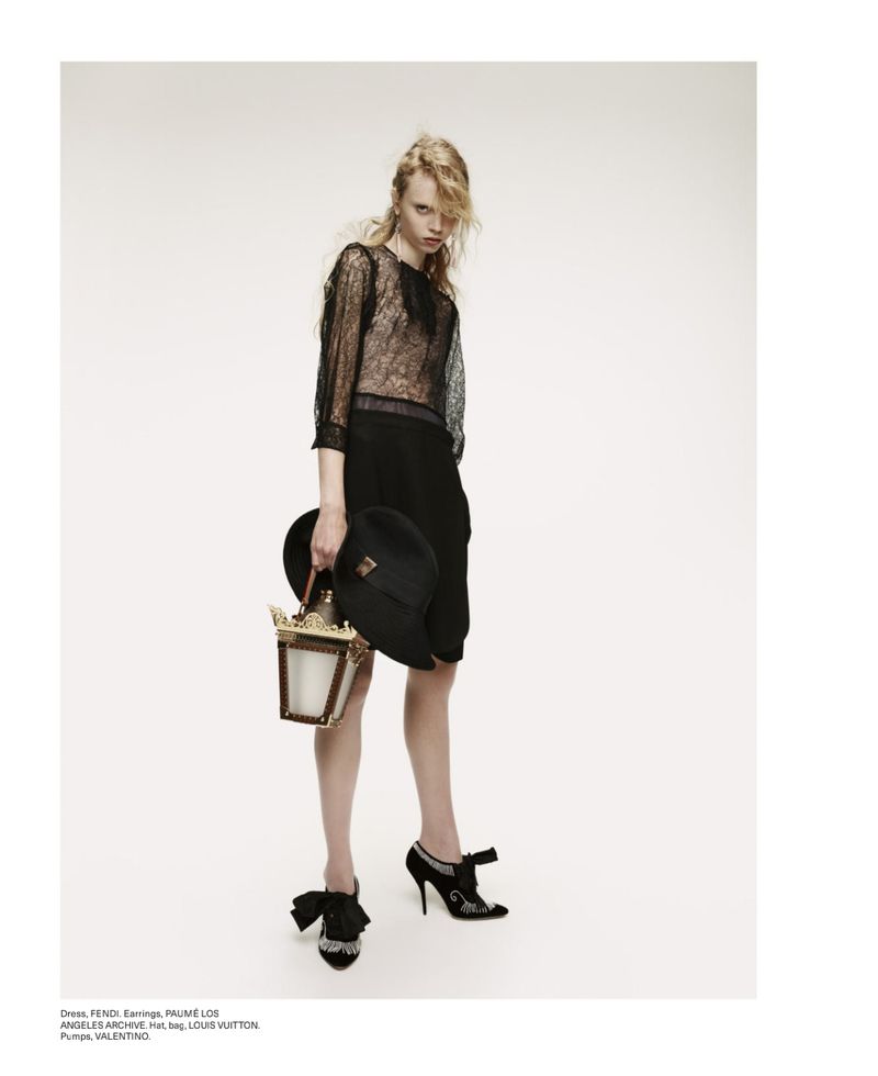 Fashion editorial shot of a slender young adult woman in a black lace top and black skirt, holding a hat and lantern.
