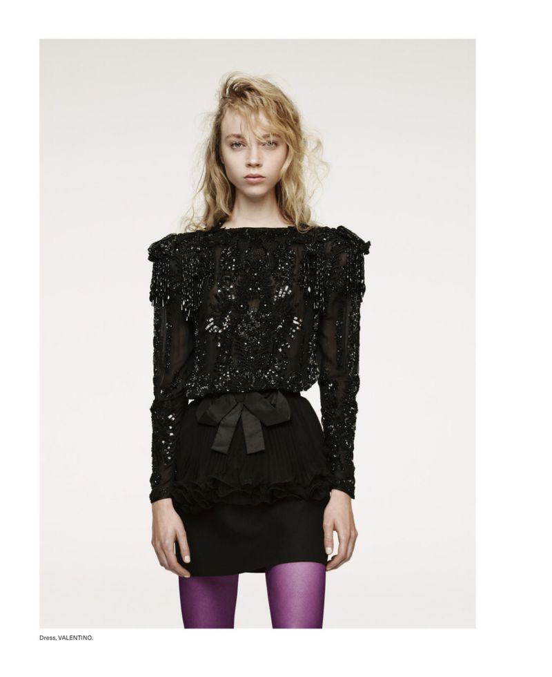 Fashion shot of a blonde woman wearing a long-sleeve embellished black top with a bow, a black skirt, and purple tights against a white background.