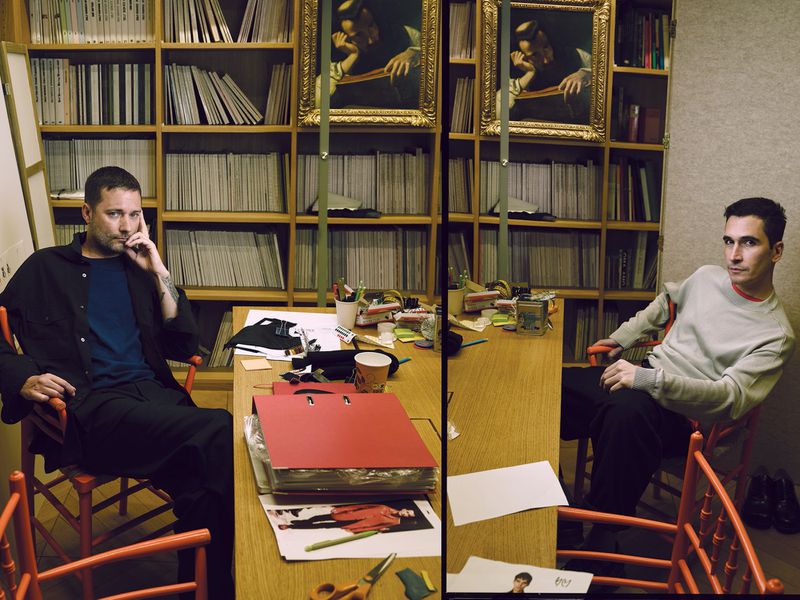 Two adult men seated at a desk in an indoor setting with bookshelves, artwork, and office supplies, creating an atmosphere of creative work.