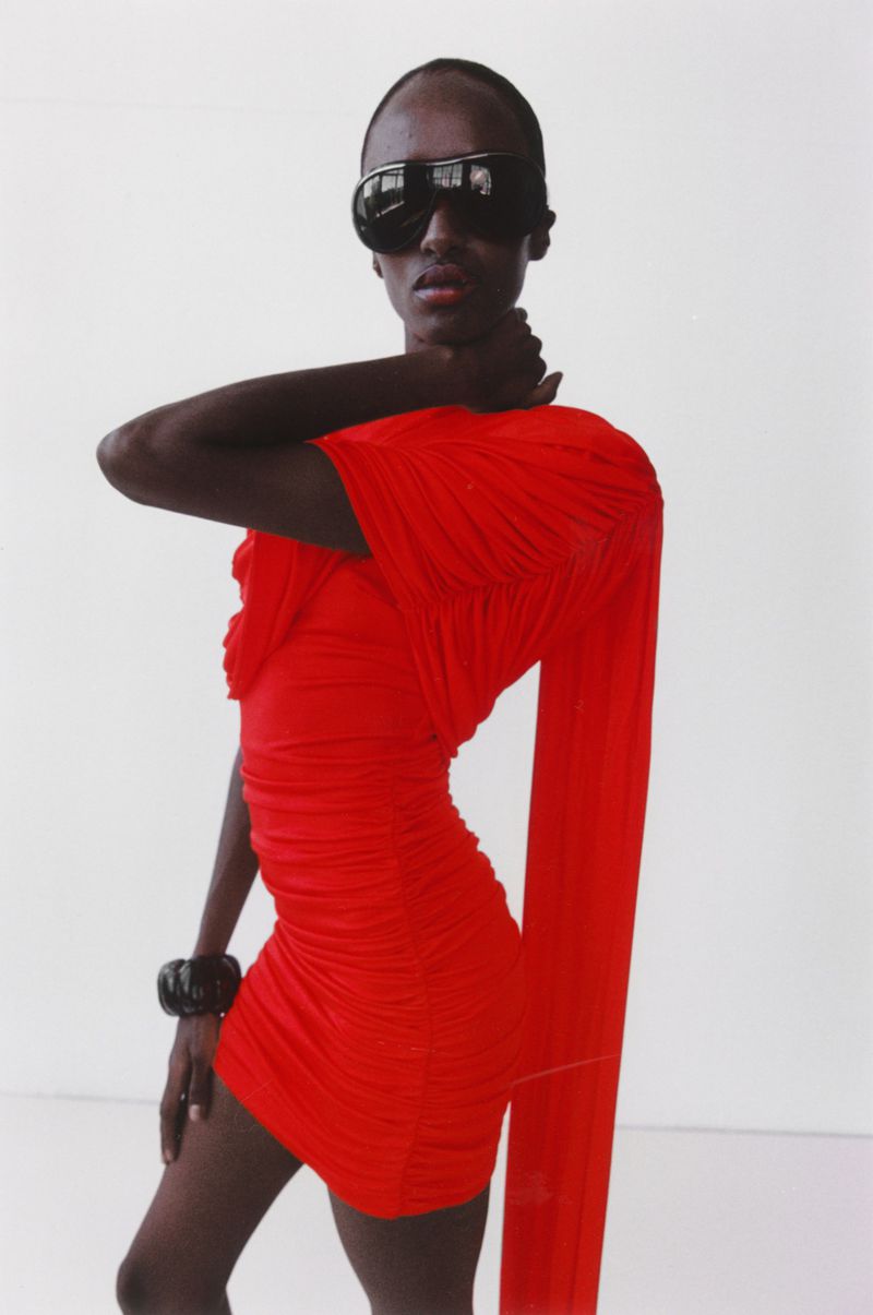 A striking fashion portrait features a Black model in a red mini dress and large sunglasses against a clean white backdrop in a studio.