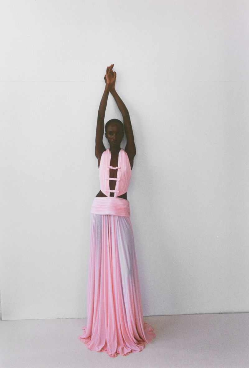 A slender dark-skinned woman in a flowing pink halter dress with her arms raised against a white wall in a fashion shot.