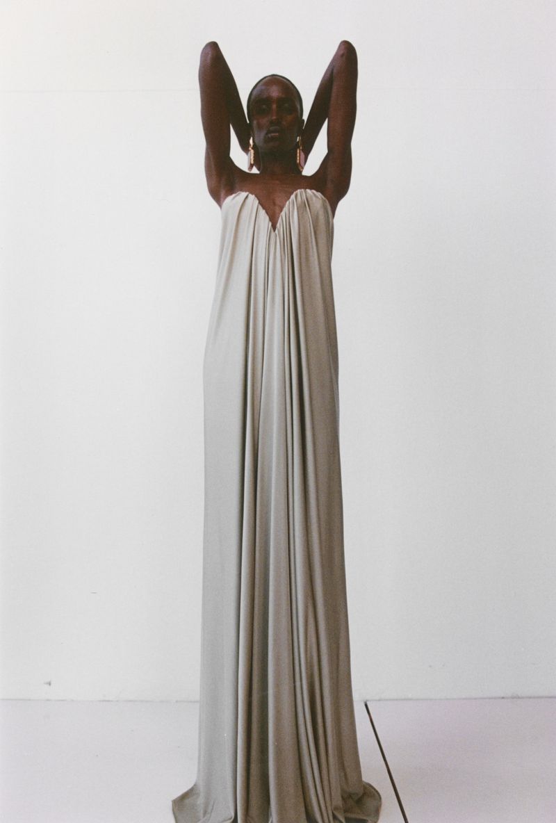 Model wearing a long strapless gathered dress with hands behind head against a white seamless background