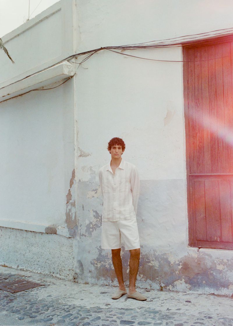 Young man in matching linen shirt and shorts poses against textured building, creating a relaxed, summery, casual fashion vibe.