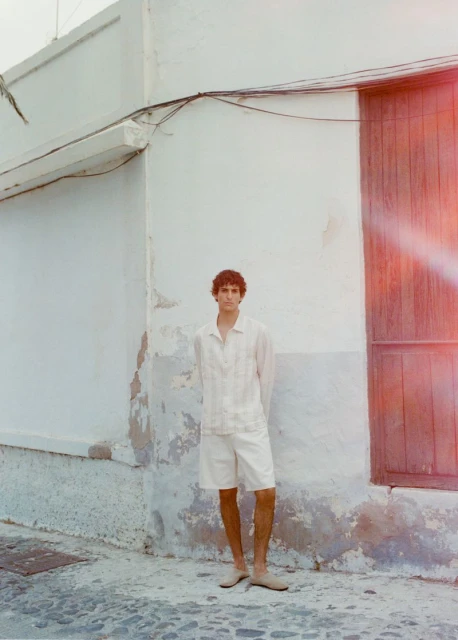 Young man in matching linen shirt and shorts poses against textured building, creating a relaxed, summery, casual fashion vibe.