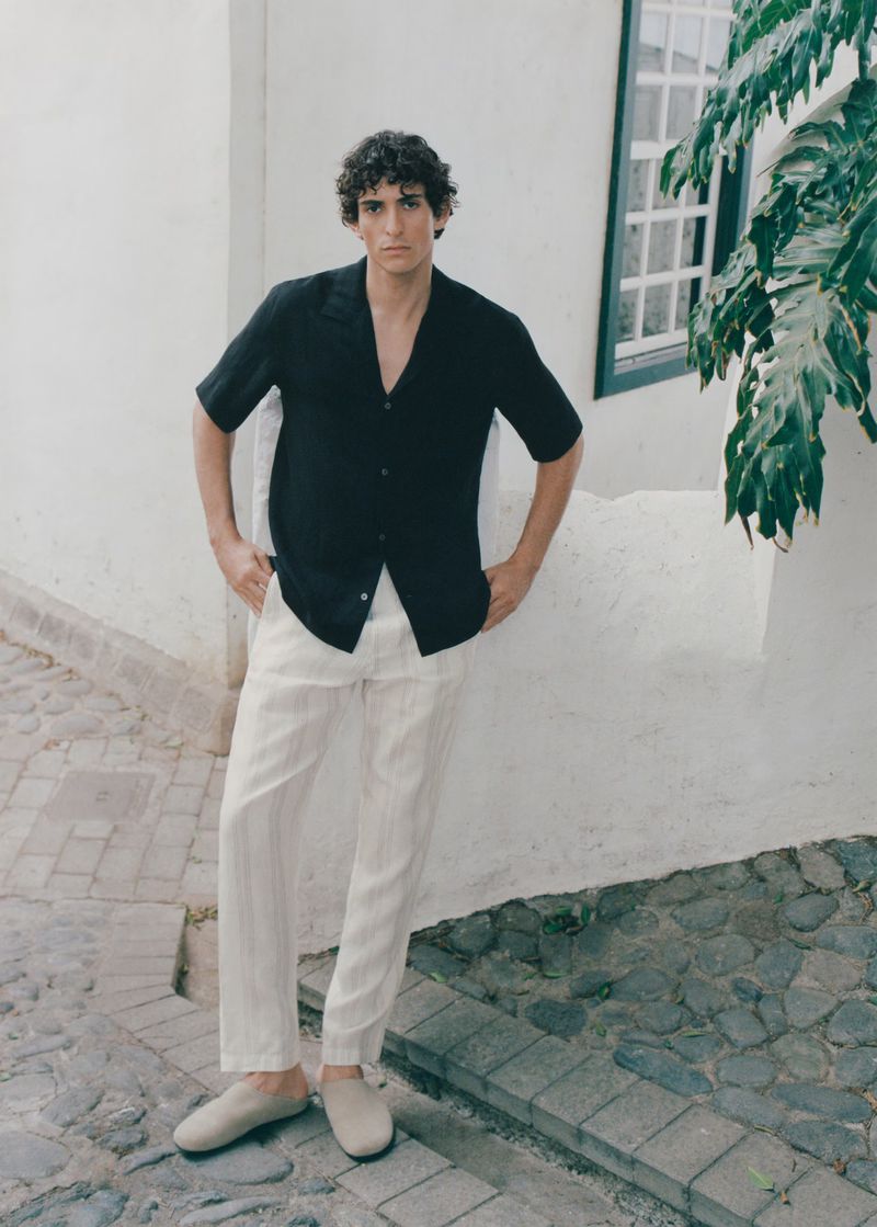 A young adult male in a dark short-sleeve button-down shirt and light pants poses against a white wall with green foliage in background.