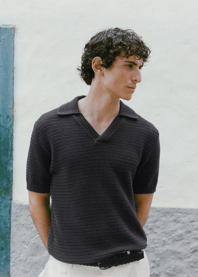 A young adult male with curly hair wearing a dark short-sleeved polo shirt and light-colored trousers stands against a textured wall.