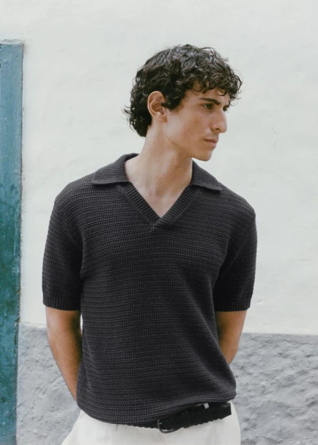 A young adult male with curly hair wearing a dark short-sleeved polo shirt and light-colored trousers stands against a textured wall.
