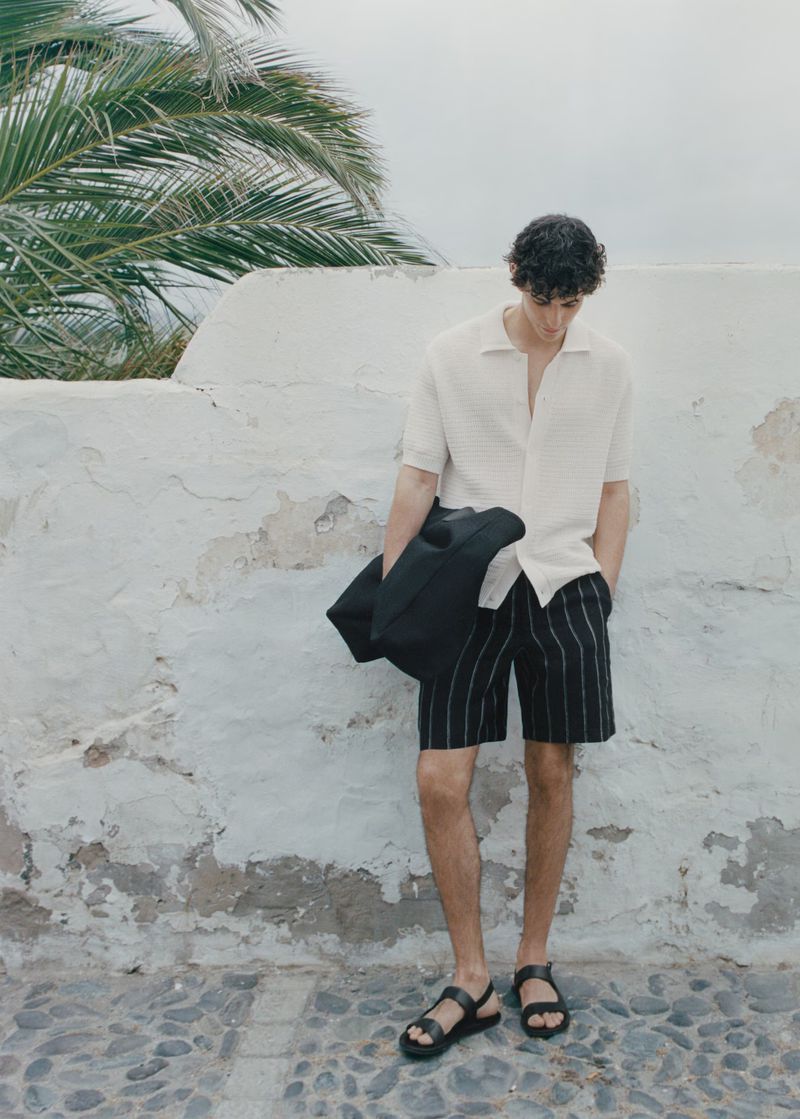 A full body shot of a man wearing a polo shirt and shorts standing near a white wall and palm tree, holding a black coat.