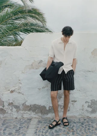 A full body shot of a man wearing a polo shirt and shorts standing near a white wall and palm tree, holding a black coat.