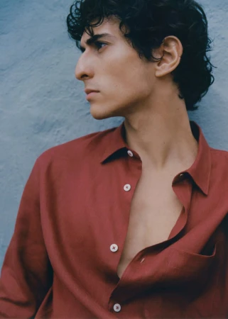 Profile view of a dark-haired man in a red button-down shirt against a textured blue background. Fashion shot, medium framing.