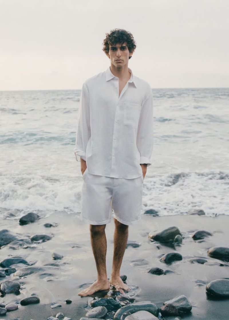 A barefoot adult male poses in a matching white linen button-down shirt and shorts on a rocky beach with ocean background.
