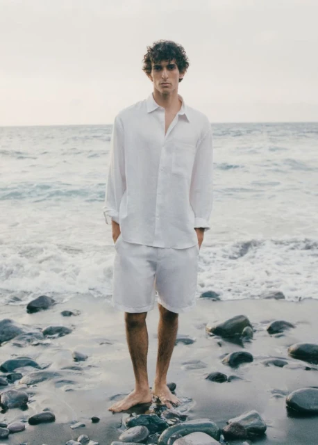 A barefoot adult male poses in a matching white linen button-down shirt and shorts on a rocky beach with ocean background.