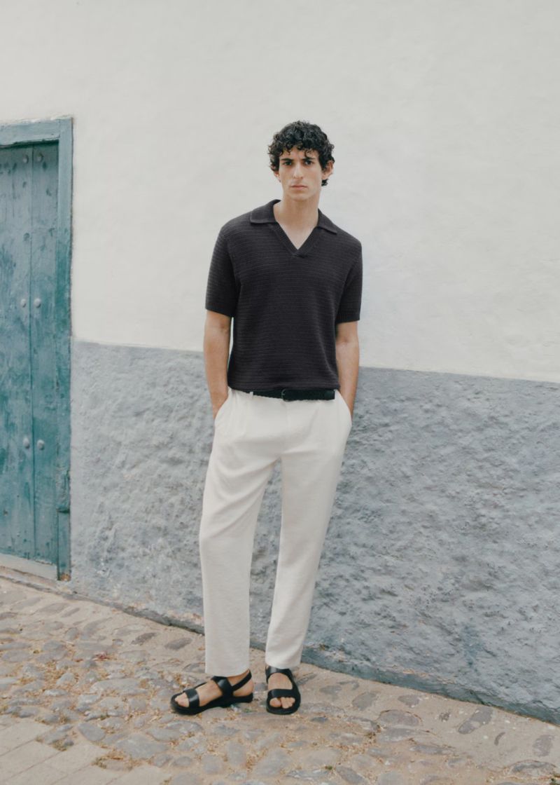 A young adult male stands against a textured wall, wearing a brown knit polo shirt, white trousers, and black sandals for a fashion editorial.