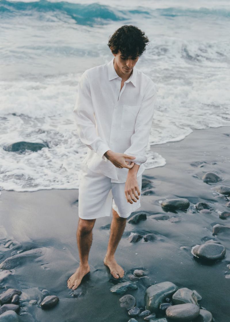 Man in white linen shirt and shorts stands barefoot on a beach with a rocky shoreline and ocean waves, adjusting his sleeve.