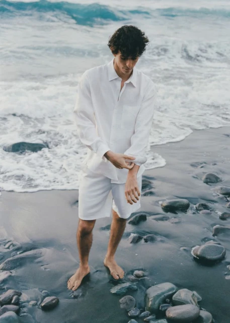 Man in white linen shirt and shorts stands barefoot on a beach with a rocky shoreline and ocean waves, adjusting his sleeve.