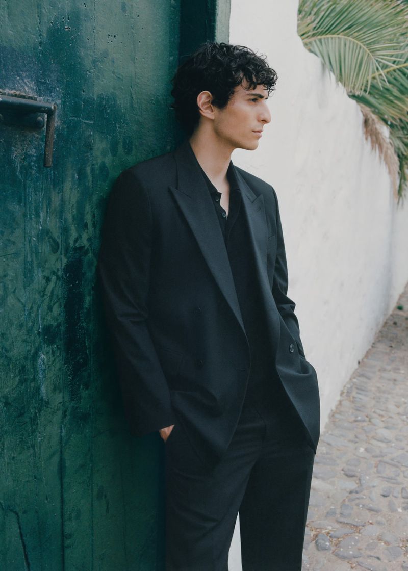 Man in a black suit jacket posing against a turquoise door outdoors, with a palm frond in the background.