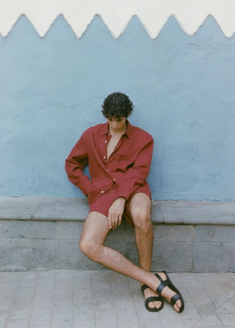 An adult male wearing a red shirt and shorts sits on a grey ledge against a light blue wall with white trim, posing in black sandals.
