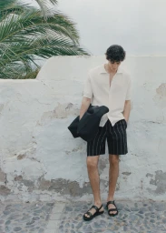 Man wearing a cream knit polo shirt and striped shorts leaning against a weathered stone wall holding a dark garment