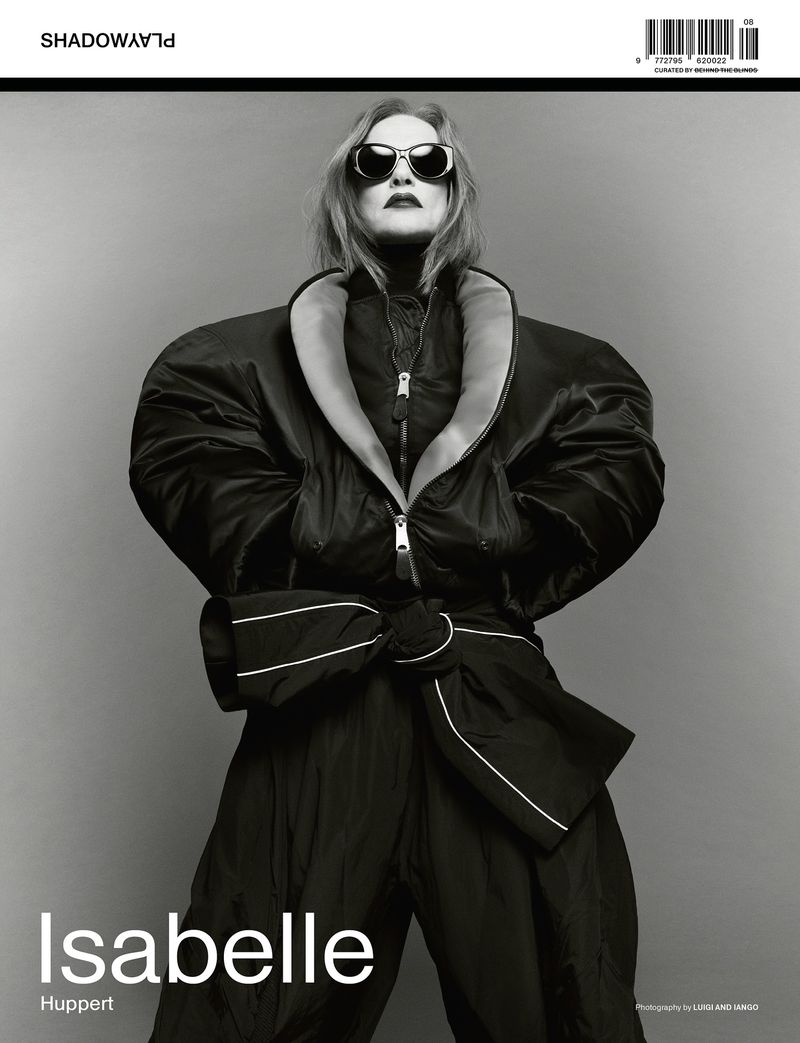 Black and white portrait of Isabelle Huppert wearing a puffy coat and sunglasses against a grey background. Magazine cover.
