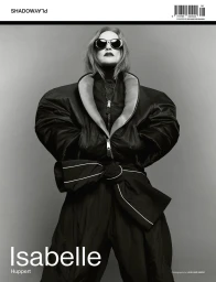 Isabelle Huppert wearing oversized black puffer jacket and sunglasses against a plain grey studio background for SHADOWPLAY Magazine.