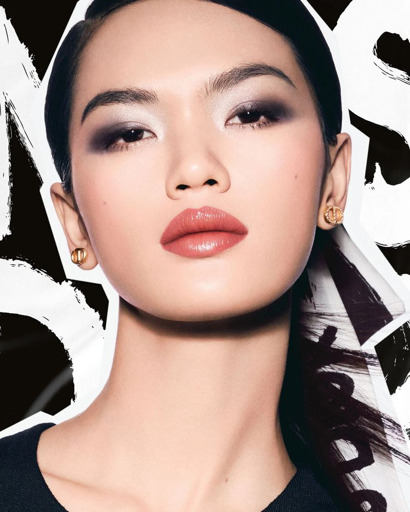 Close-up portrait of a beautiful Asian woman with smoky eyeshadow, glossy lips, gold earrings, and a blurred black and white background.