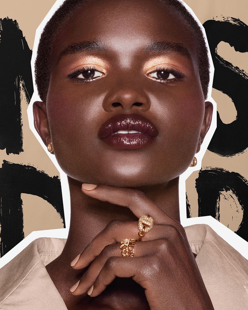 Close-up beauty shot of a black female model with glossy dark lips, gold eyeshadow, and statement rings against a patterned backdrop.