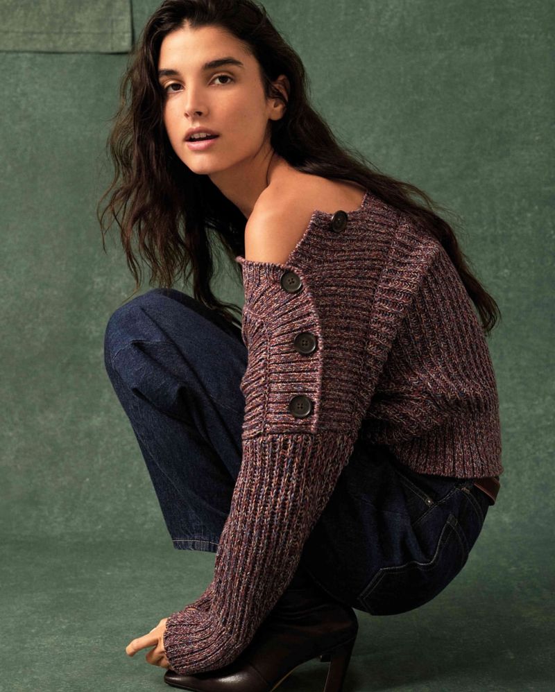 Model crouching in a ribbed off-the-shoulder plum knit sweater with button details and dark denim jeans against a green textured backdrop.