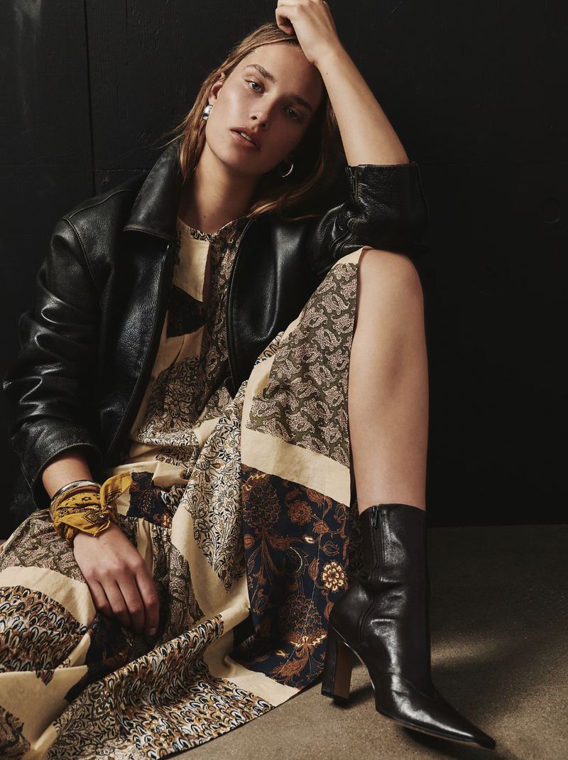 A woman in a patterned dress, leather jacket, and boots poses in a studio, casting a chic and confident figure against a dark background.