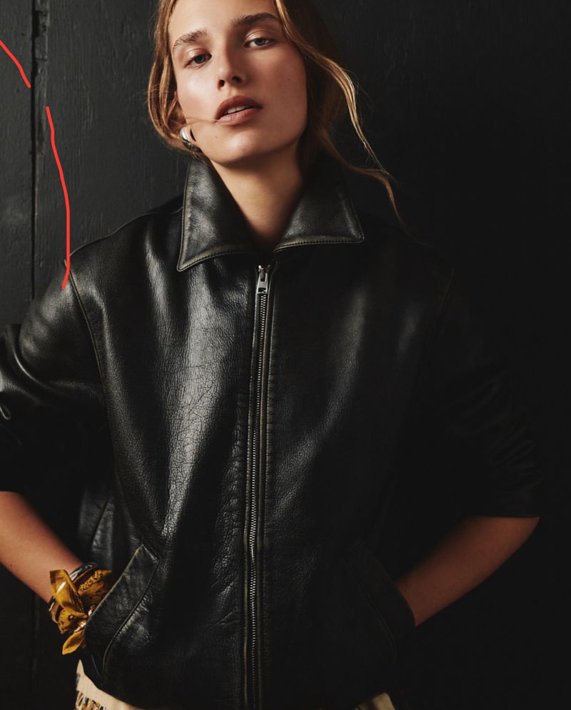 A blonde woman in a black leather jacket poses indoors against a dark background, looking directly at the camera with a neutral expression.