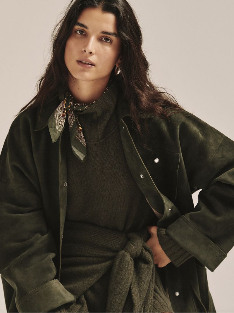 A young woman with long dark hair wears a dark green sweater dress with a knotted belt and a matching jacket in a studio shot.