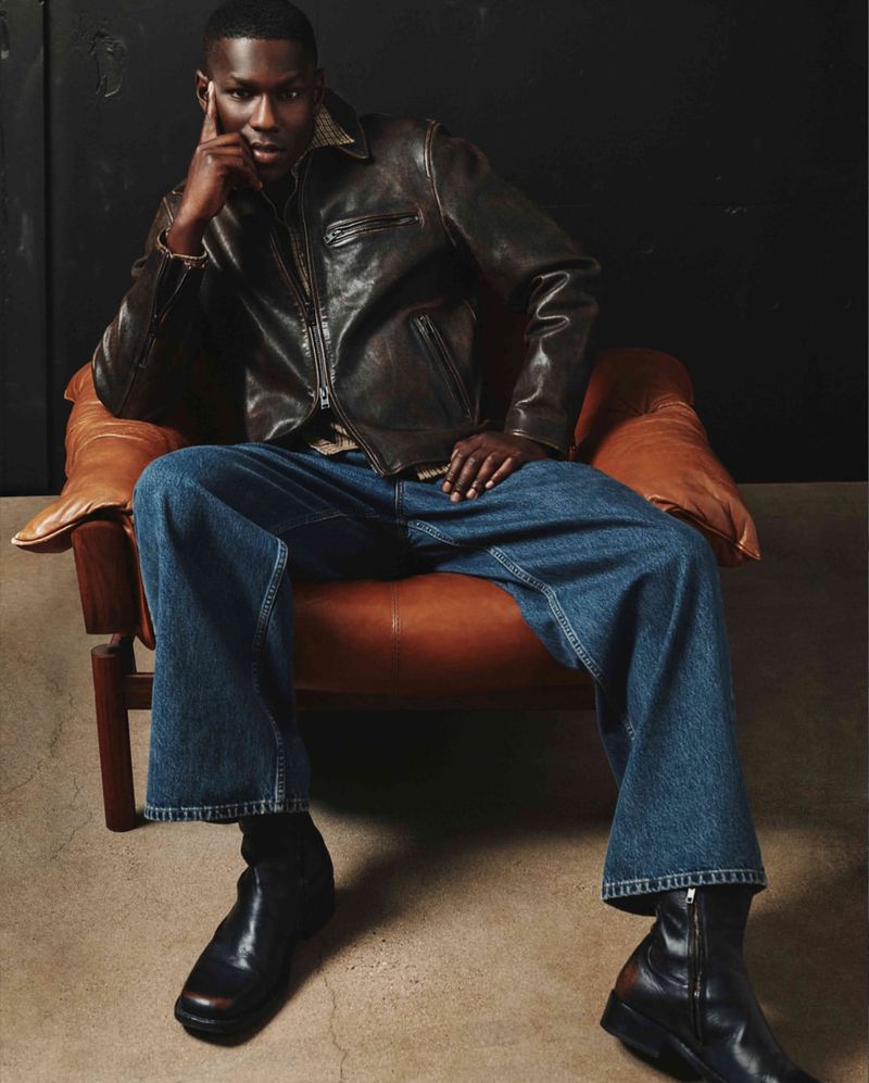 A dark-skinned male model is seated in an armchair, wearing a brown leather jacket, wide-leg blue jeans, and black boots against a dark backdrop.