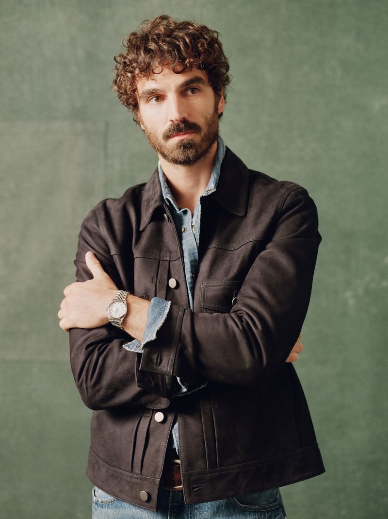 Portrait of a man with curly hair and a beard wearing a brown jacket and a blue shirt standing in front of a green background.