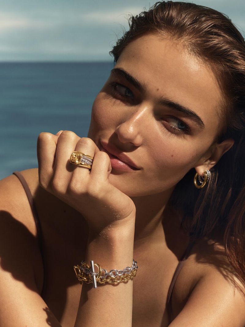 Woman poses outdoors with gold ring and bracelet jewelry, ocean backdrop, natural light, medium shot, accessorized fashion.