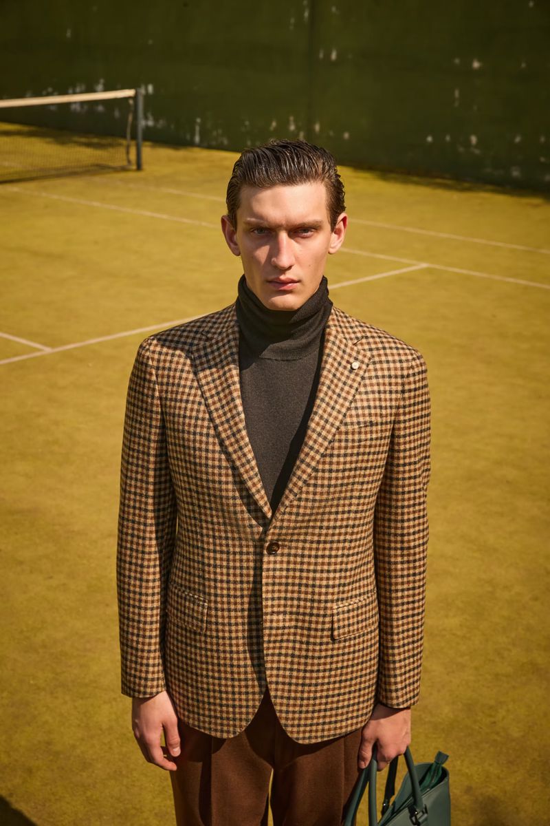 Man in patterned blazer and turtleneck stands on tennis court holding green bag, looking directly at camera in a medium shot.