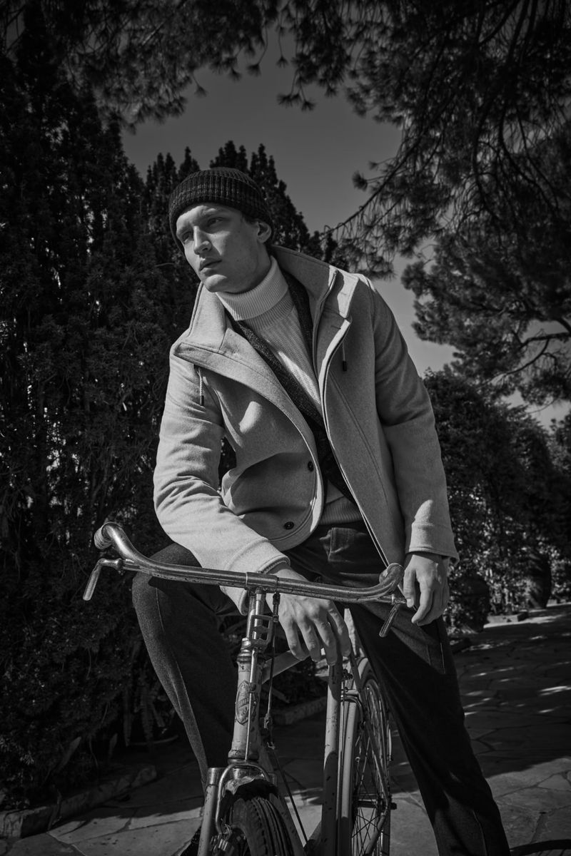 Monochrome portrait of a stylish man wearing a coat and beanie, posed with a vintage bicycle in a sunlit park setting.