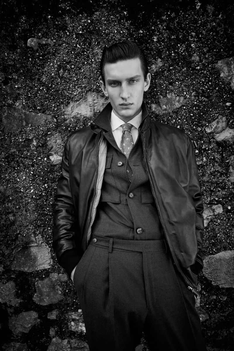 Monochrome portrait of a serious young man in a jacket and suit posing against a textured wall, evoking a vintage aesthetic.