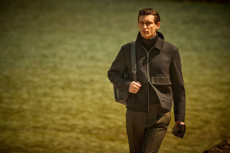 A stylish man in a black jacket and turtleneck poses outdoors with a messenger bag, against a blurred water background in an outdoor fashion shot.