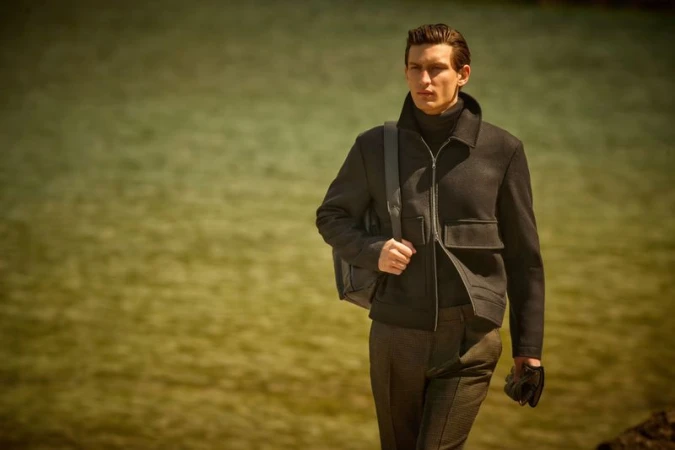 A stylish man in a black jacket and turtleneck poses outdoors with a messenger bag, against a blurred water background in an outdoor fashion shot.