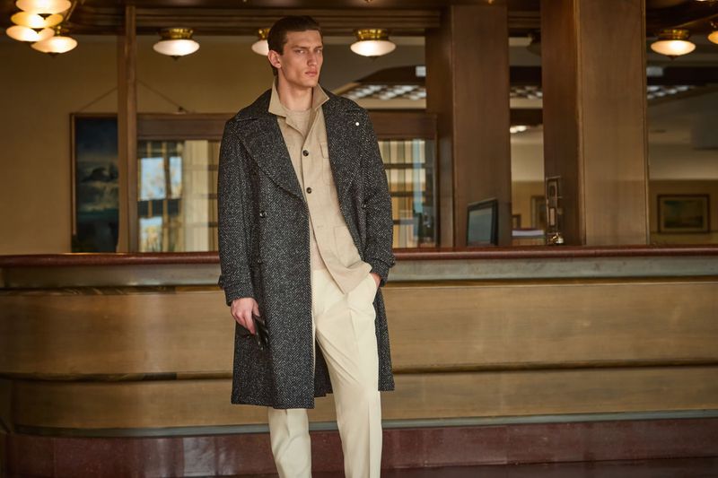 Fashion shot of a male model posing in front of a classic hotel lobby. He wears a stylish grey coat over a beige shirt and pants.