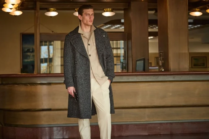 Fashion shot of a male model posing in front of a classic hotel lobby. He wears a stylish grey coat over a beige shirt and pants.