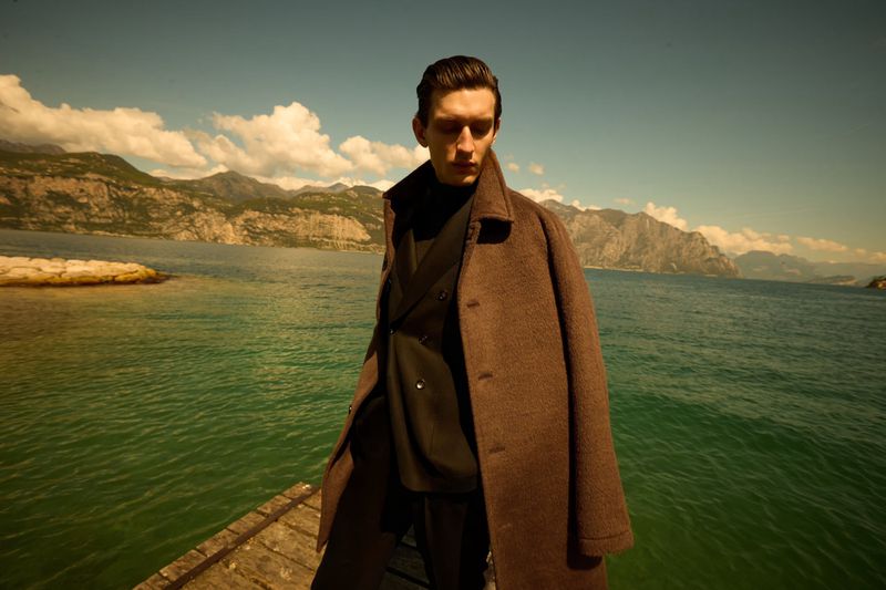 Male model in brown coat poses on wooden dock with lake and mountains backdrop on a sunny day for fashion editorial content.