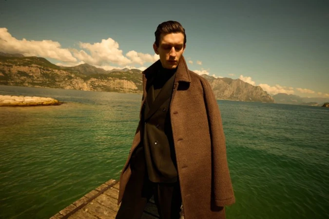 Male model in brown coat poses on wooden dock with lake and mountains backdrop on a sunny day for fashion editorial content.