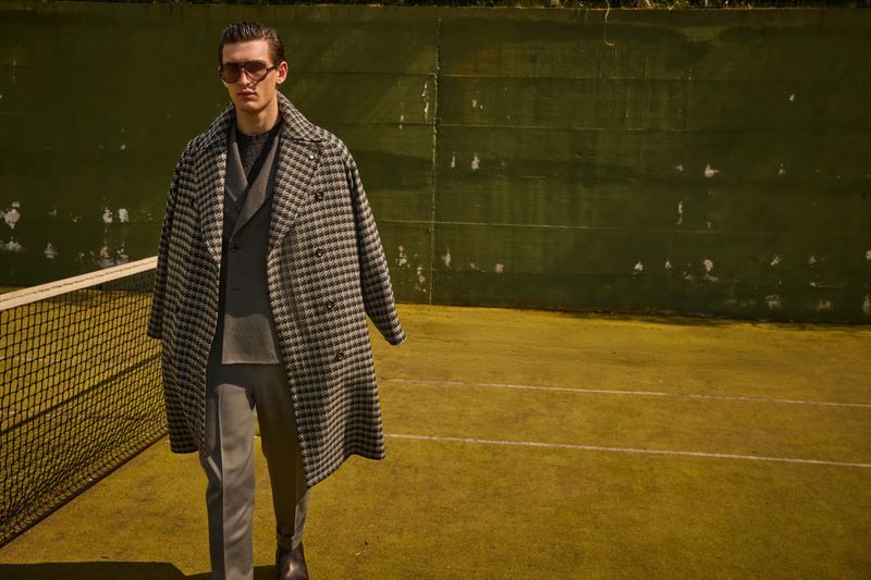 A fashionable adult male walks on a tennis court wearing a grey suit, black boots, sunglasses and a black and white checkered overcoat.