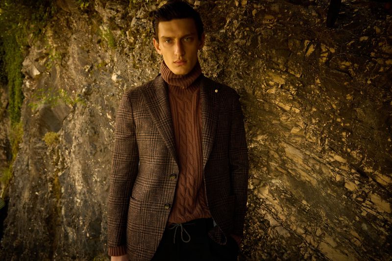 Adult male poses in front of a rock wall, wearing a brown blazer, turtleneck sweater, and black pants for fashion editorial content.